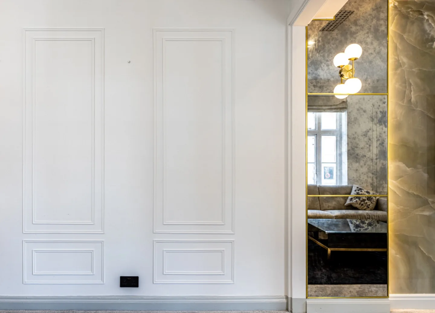 Classic white decorative wall panelling beside a brass-framed antiqued mirror and onyx-clad doorway