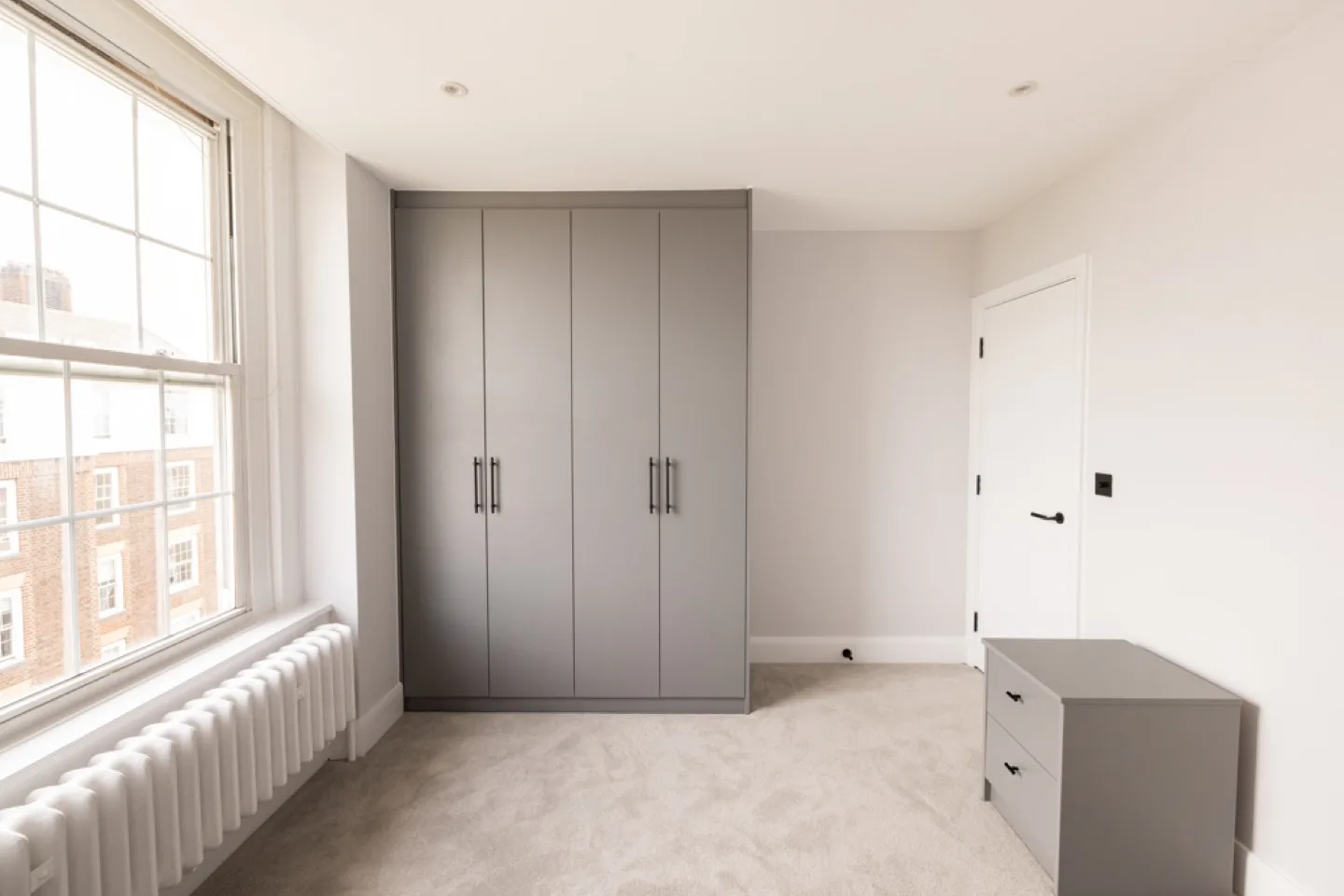 Grey four-door fitted wardrobe and matching bedside chest in a bright bedroom by a sash window
