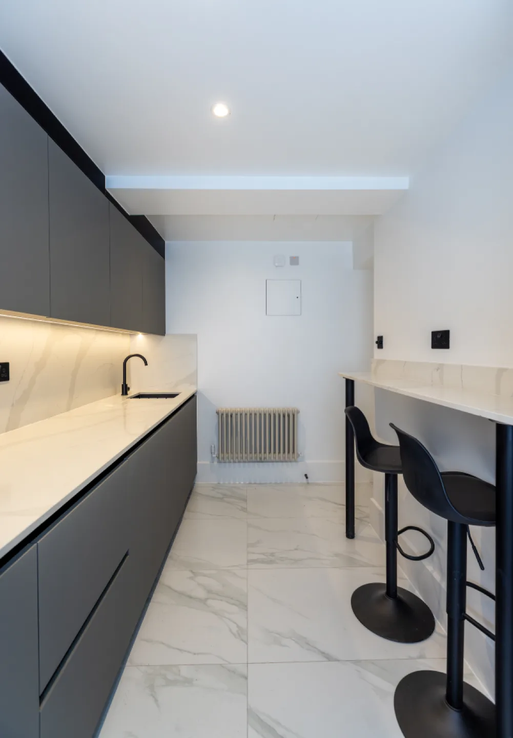 Galley kitchen with grey handleless cabinets, white quartz worktop, black tap and breakfast bar with stools