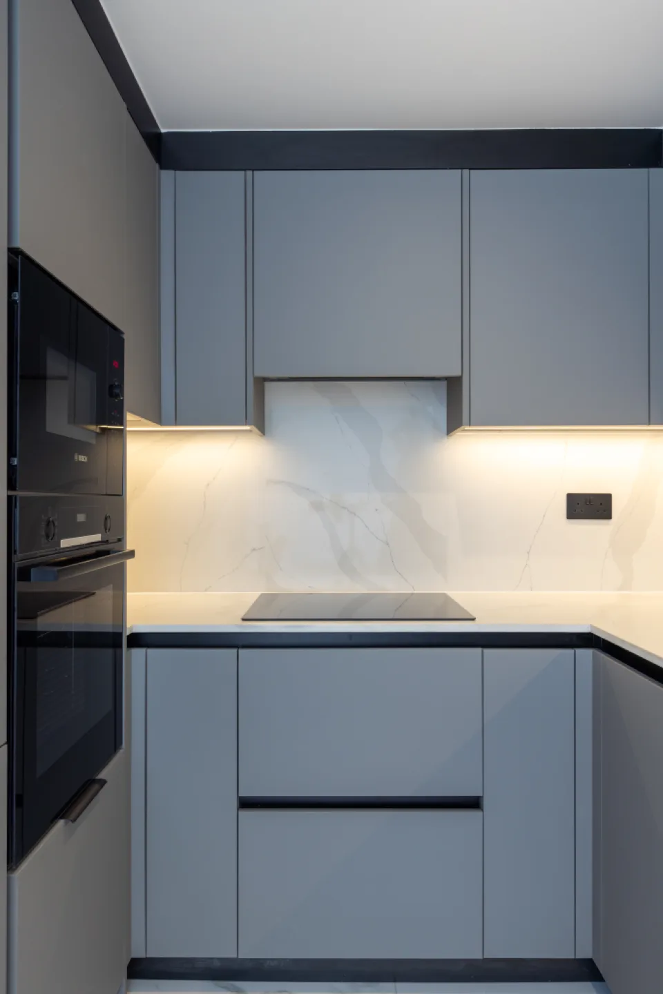 Grey handleless cabinets with black profiles, induction hob and marble splashback below illuminated wall units