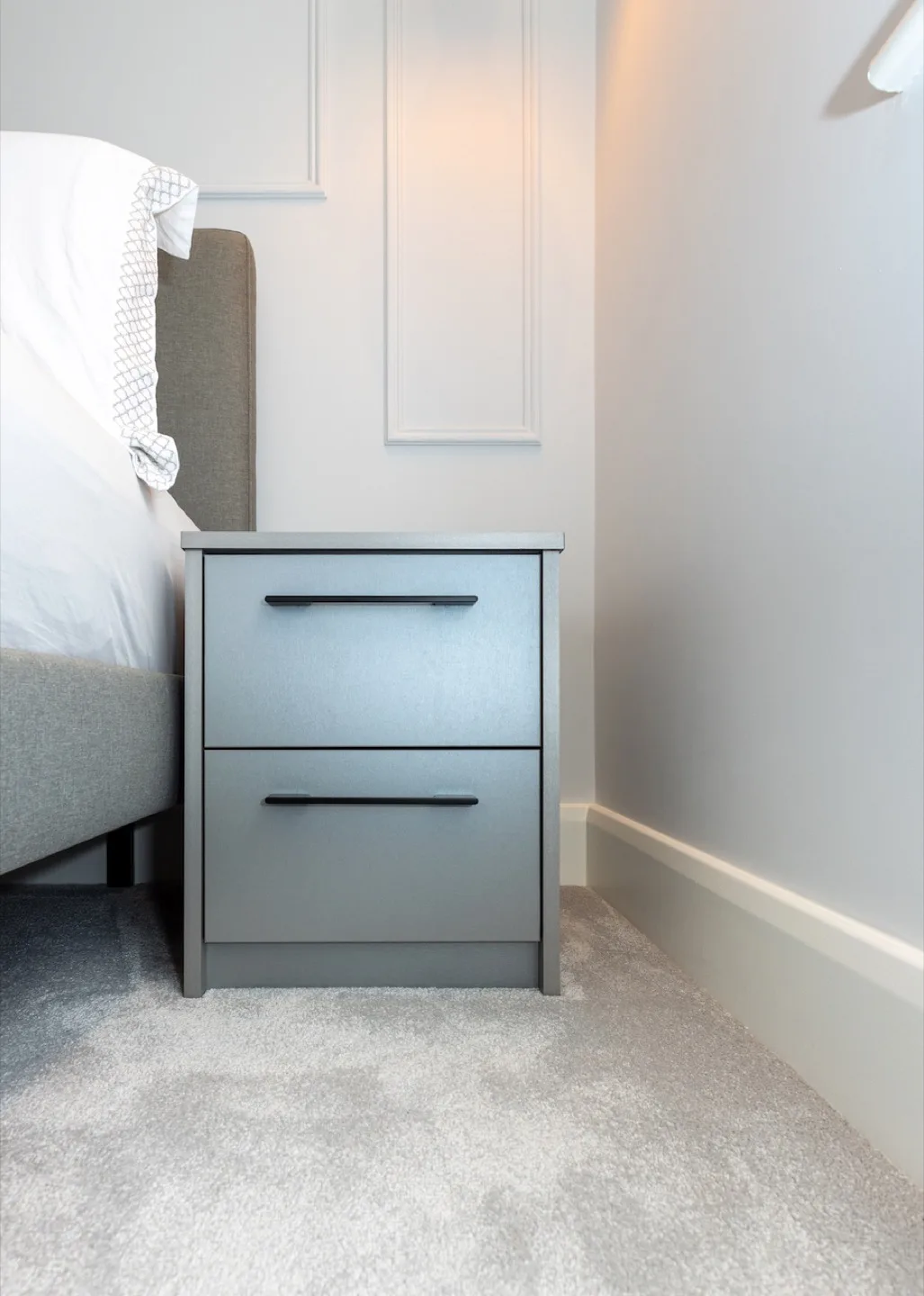 Bespoke grey two-drawer bedside table with black bar handles beside the bed, against panelled walls