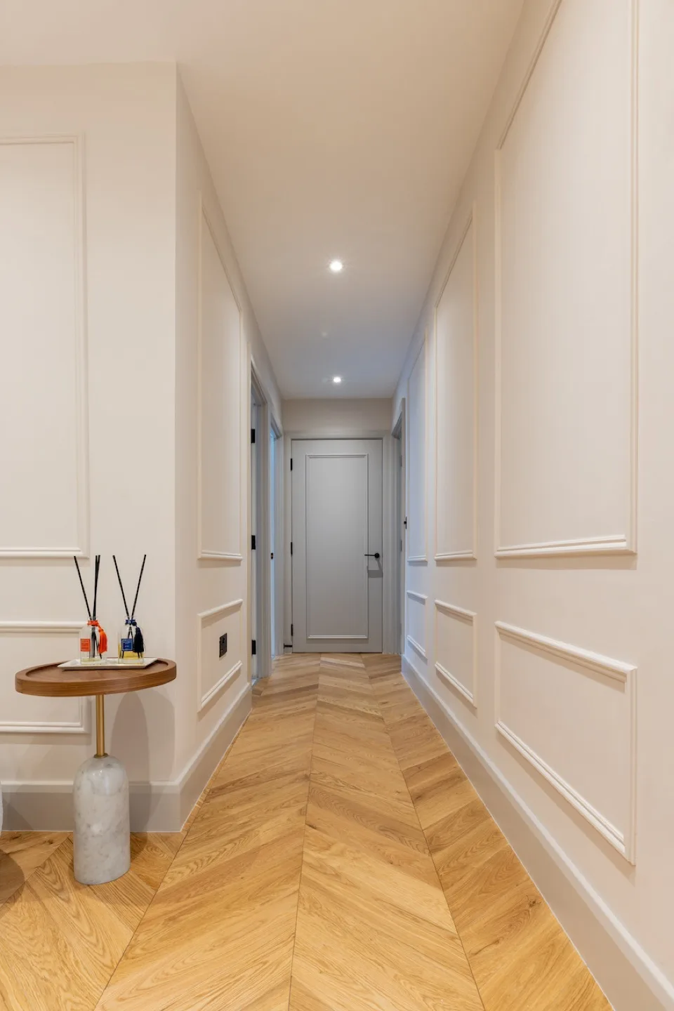 Hallway lined with modern white French panelling and herringbone oak flooring leading to a grey door