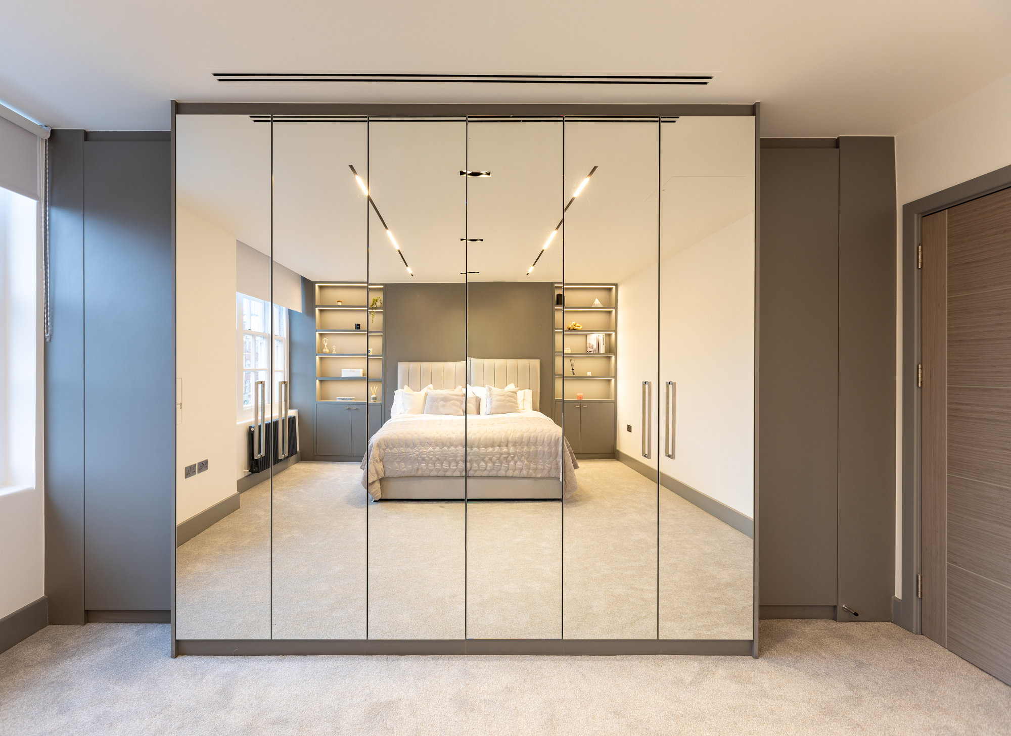Floor-to-ceiling bespoke fitted mirrored wardrobe doors with LED display niches on either side in a Kensington bedroom