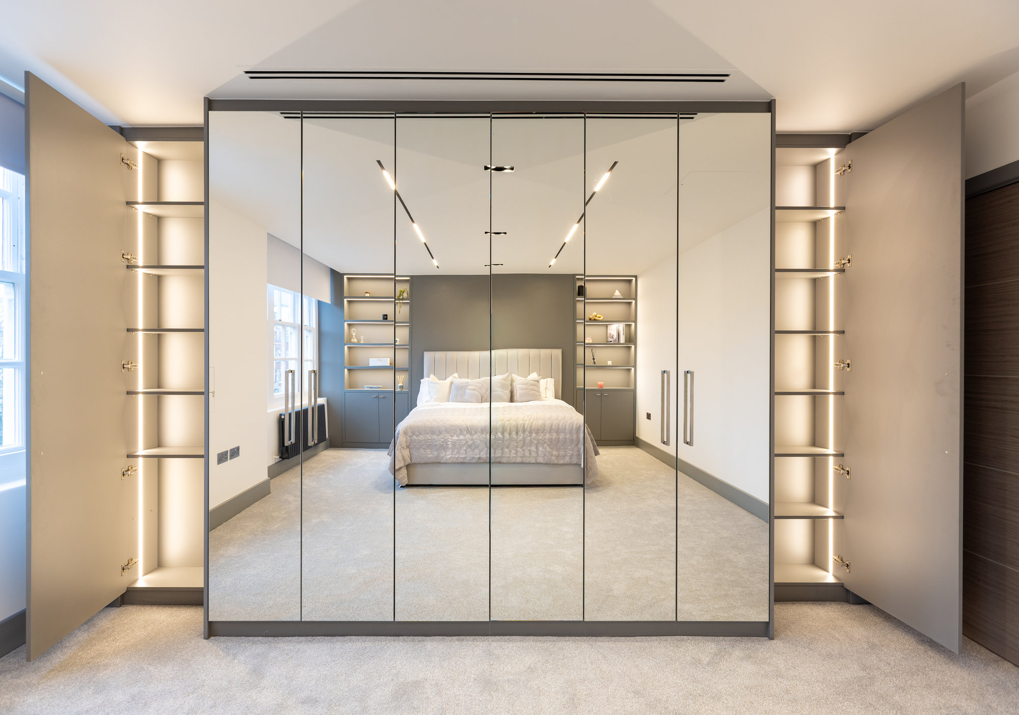 Full-wall bespoke fitted mirrored wardrobe flanked by LED-lit open display shelves in a luxurious Kensington bedroom
