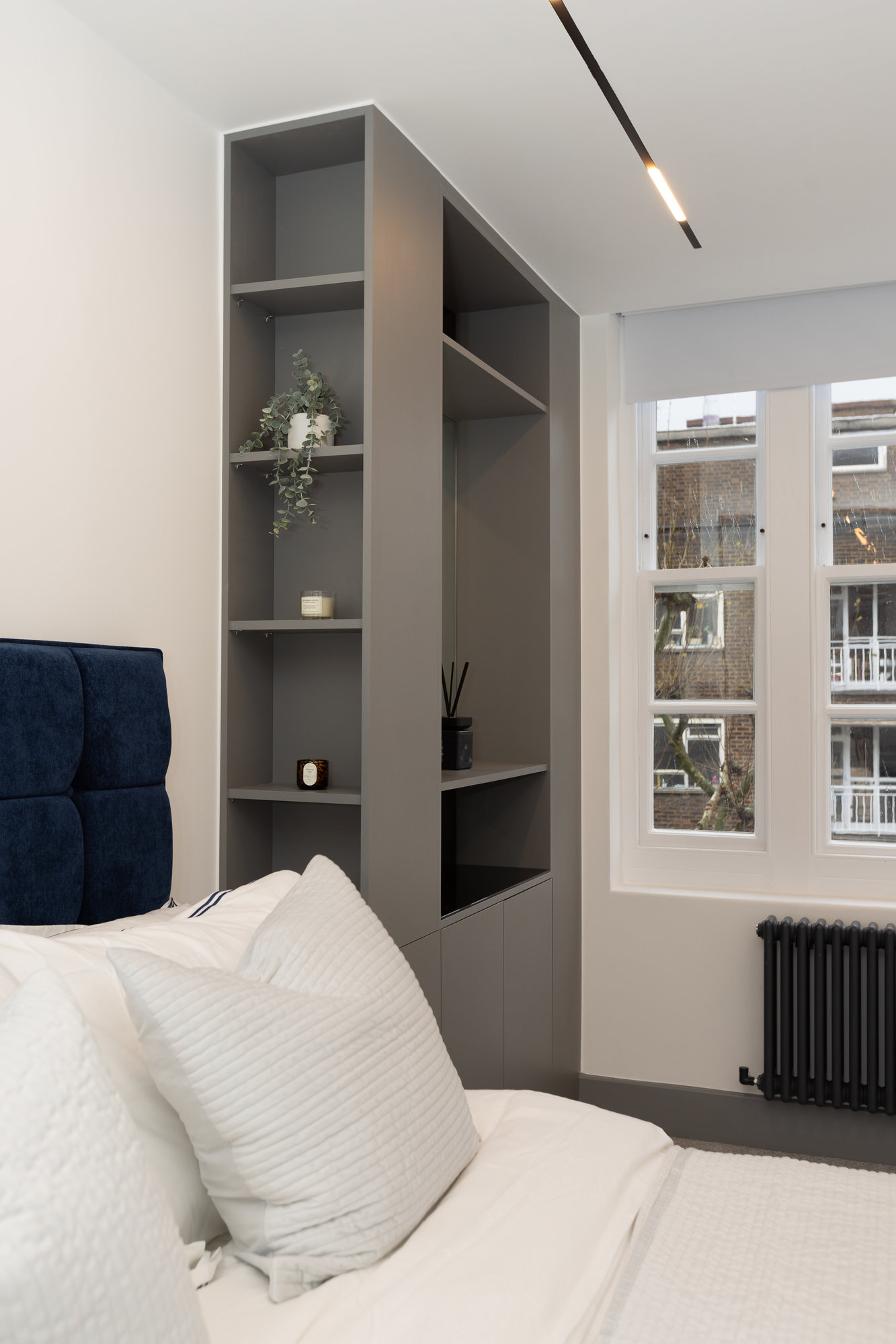 Grey bespoke fitted display shelving unit with open shelves styled with decorative items beside a navy upholstered bed in a Kensington bedroom