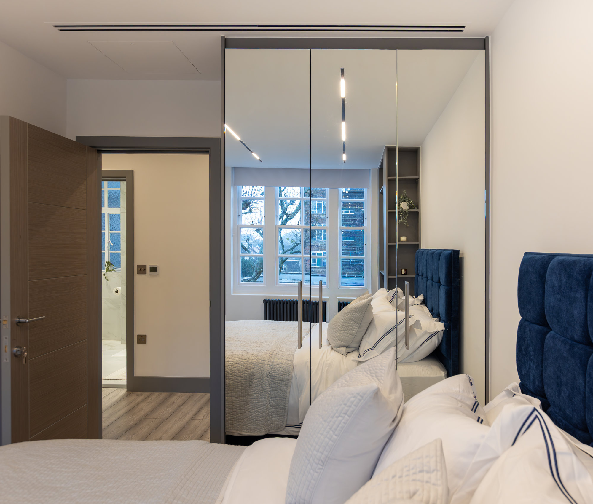 Four-door bespoke fitted wardrobe with full-length mirrored doors reflecting a modern Kensington bedroom with navy headboard