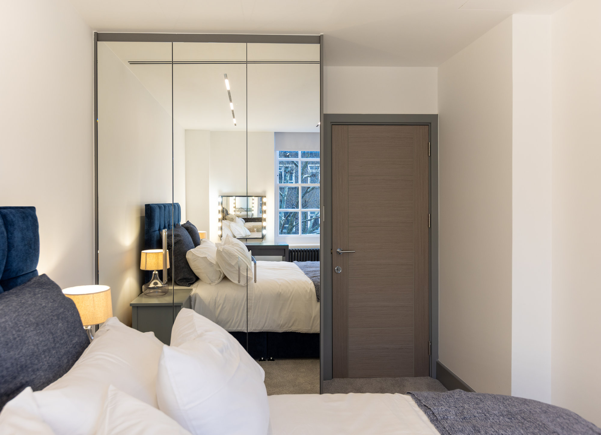 Wide-angle view of bespoke fitted mirrored wardrobe beside bedroom doorway in a contemporary Kensington apartment