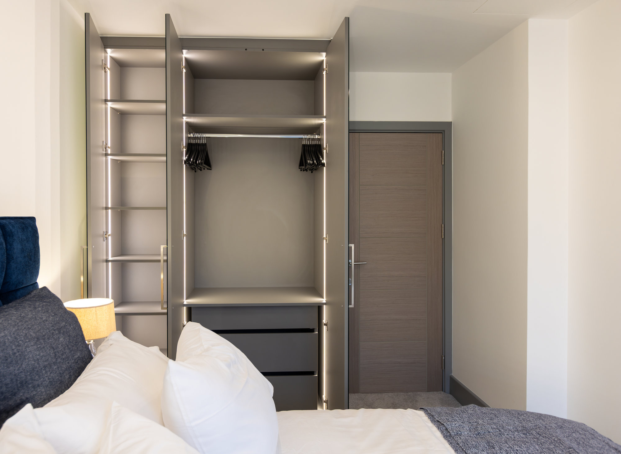 Bespoke fitted wardrobe open showing LED-lit interior with soft-close drawers and hanging rail near bedroom door in Kensington