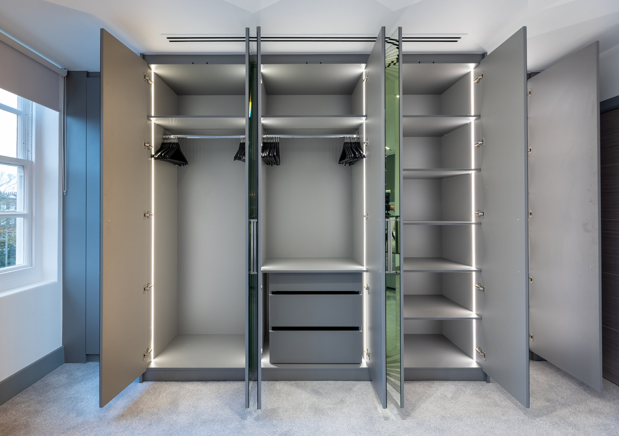 Bespoke fitted wardrobe open showing LED-lit storage with hanging rails, drawers, and central mirror panel in a Kensington home
