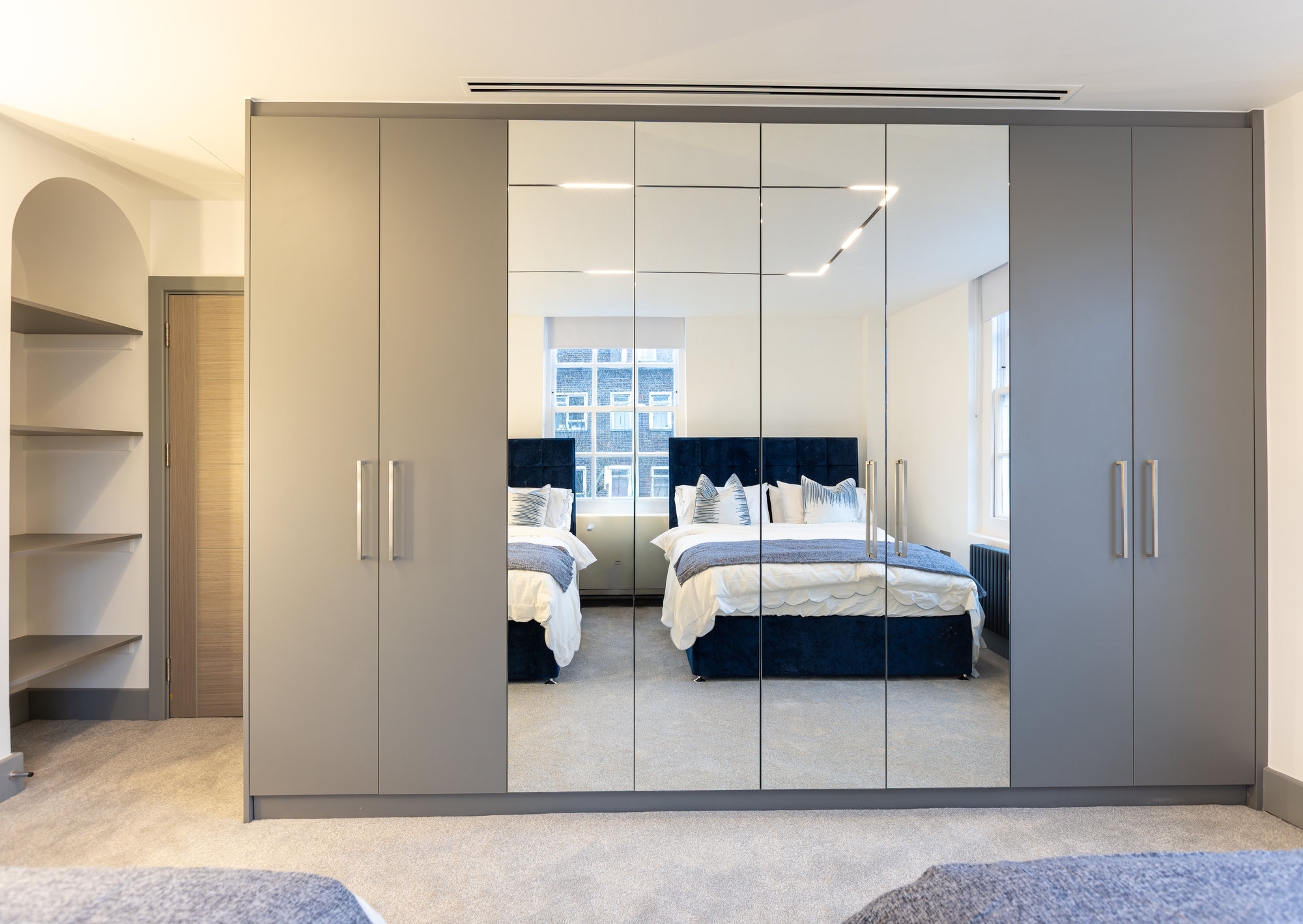 Large six-door bespoke fitted wardrobe with grey panels and central mirrored doors reflecting the bed in a Kensington master bedroom