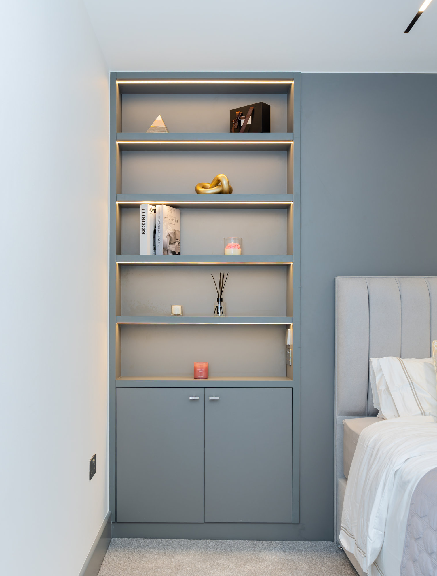Bespoke built-in bookshelf with brass-edged shelves and lower cupboard in a blue-grey bedroom wall beside the bed in Kensington