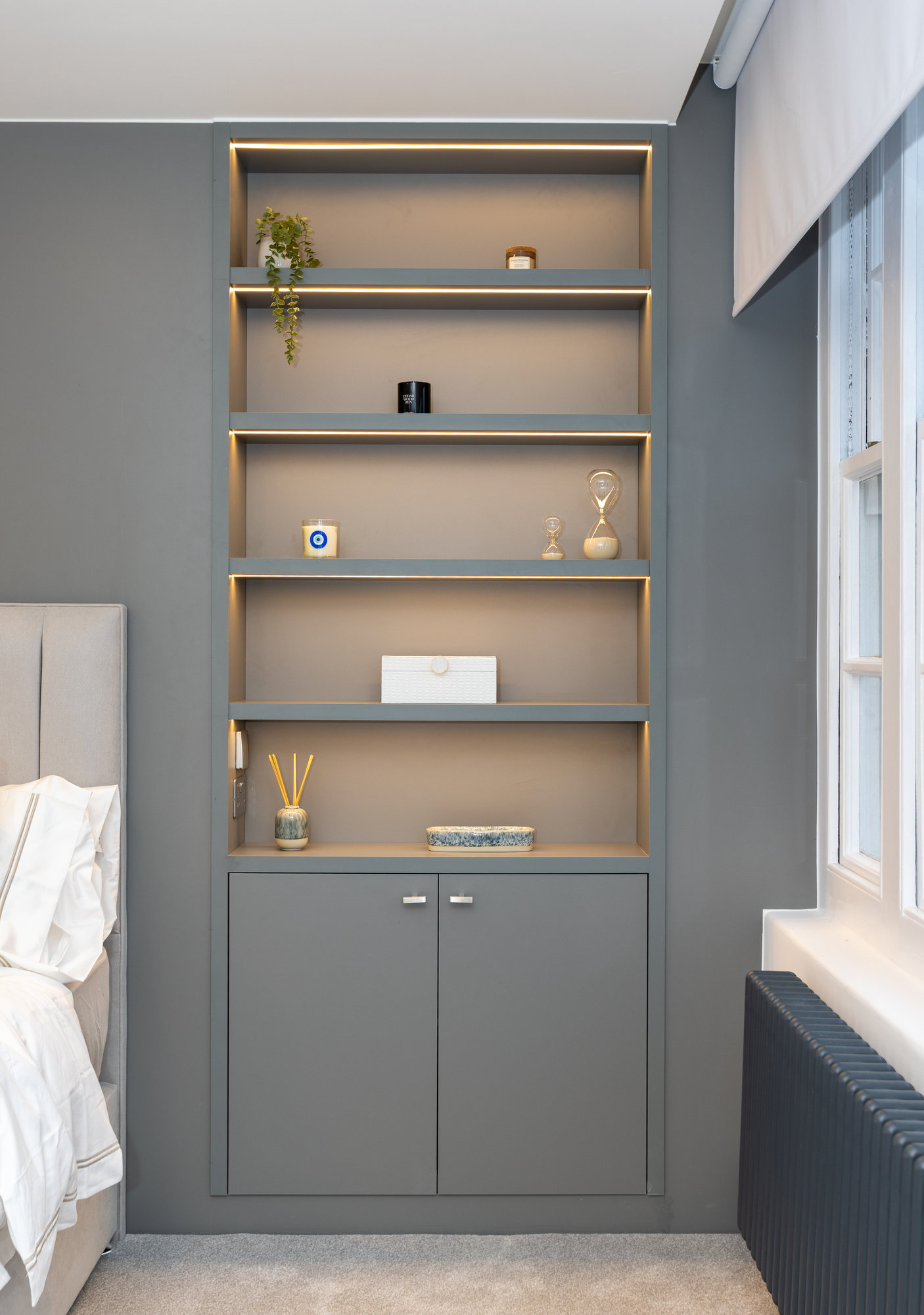 Bespoke floor-to-ceiling shelving unit with closed cupboard and backlit brass-edged shelves in a Kensington bedroom
