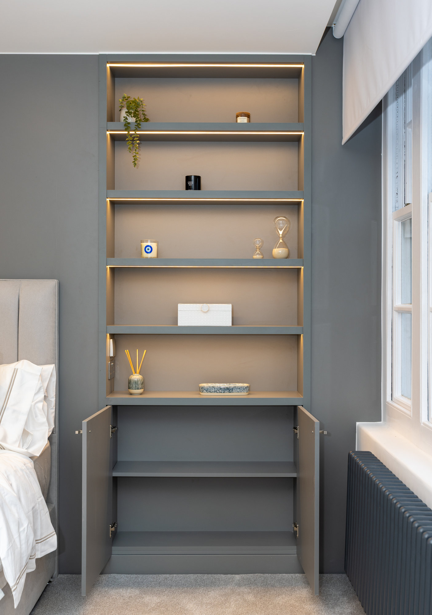 Full view of bespoke integrated shelving beside bed with open cupboard doors and brass-trimmed shelves in Kensington