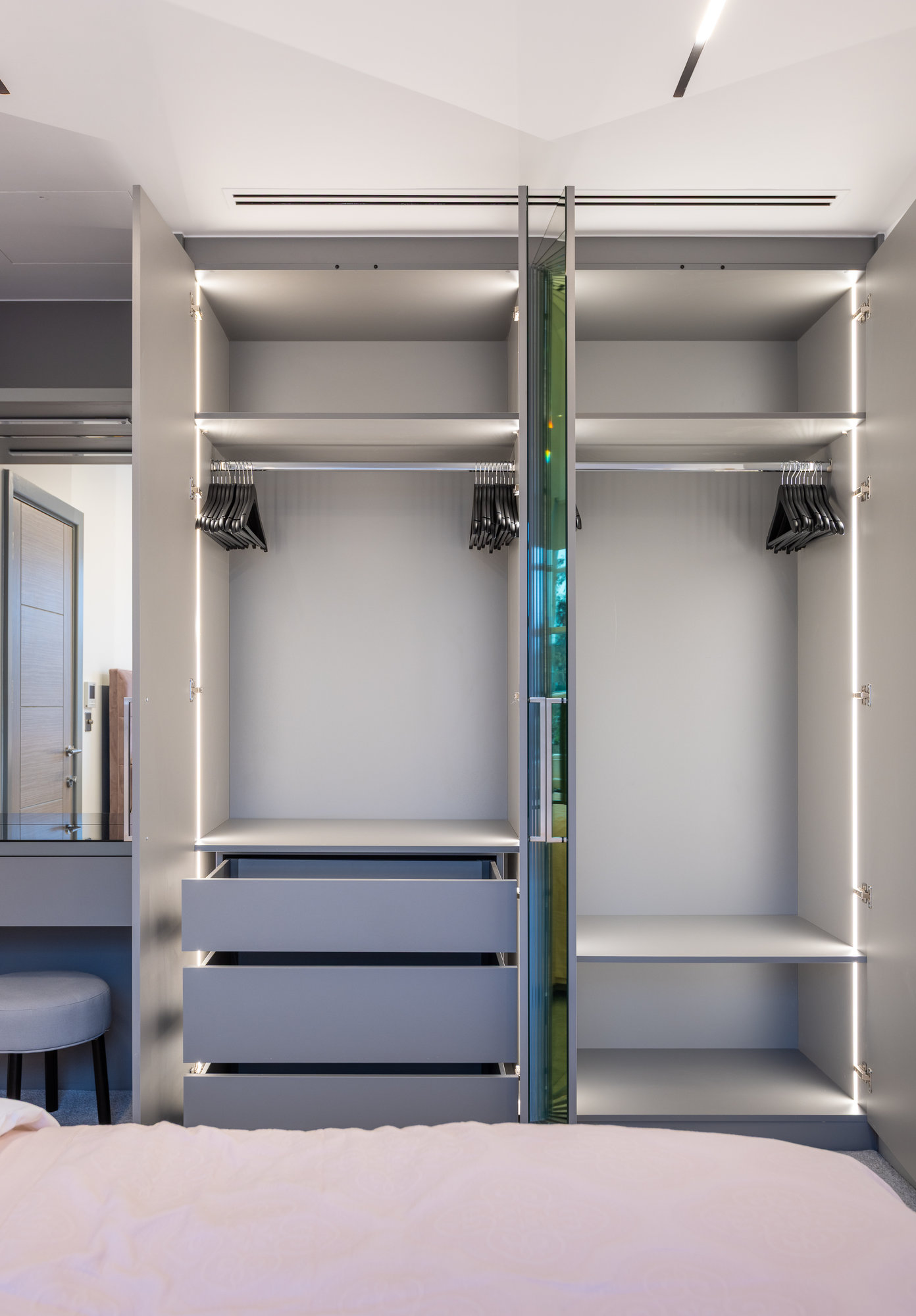 Bespoke wardrobe doors open revealing LED-illuminated interior with dual hanging rails and drawers beside a vanity desk in Kensington