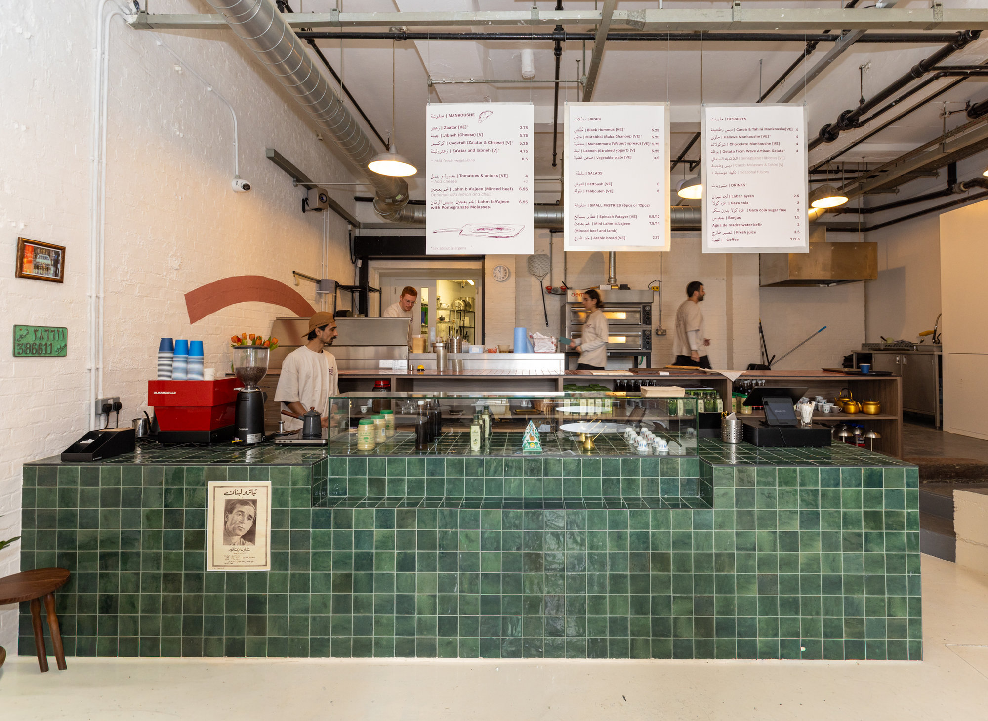 Green zellige-tiled service counter with hanging menu boards and open kitchen pass in a Dalston restaurant