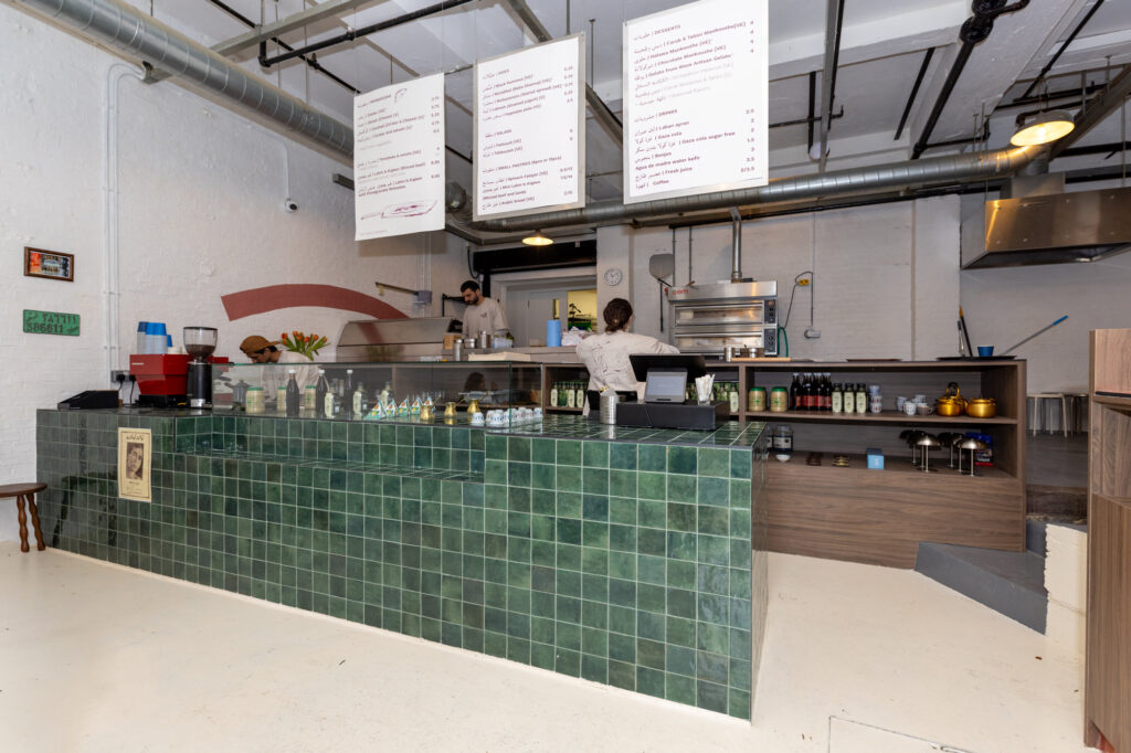 Wide-angle view of green zellige-tiled service counter with menu boards and kitchen pass in a Dalston commercial space