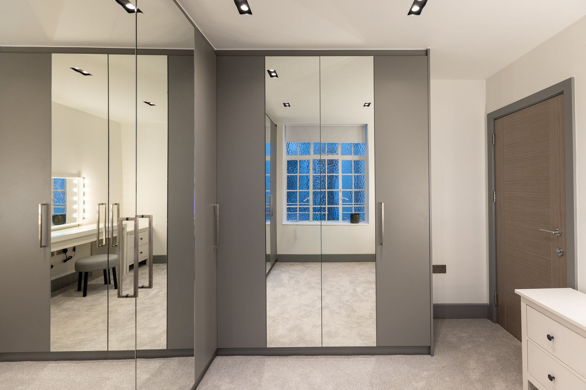 Corner view of bespoke walk-in wardrobe with mirrored and grey doors alongside a vanity with drawers in Kensington