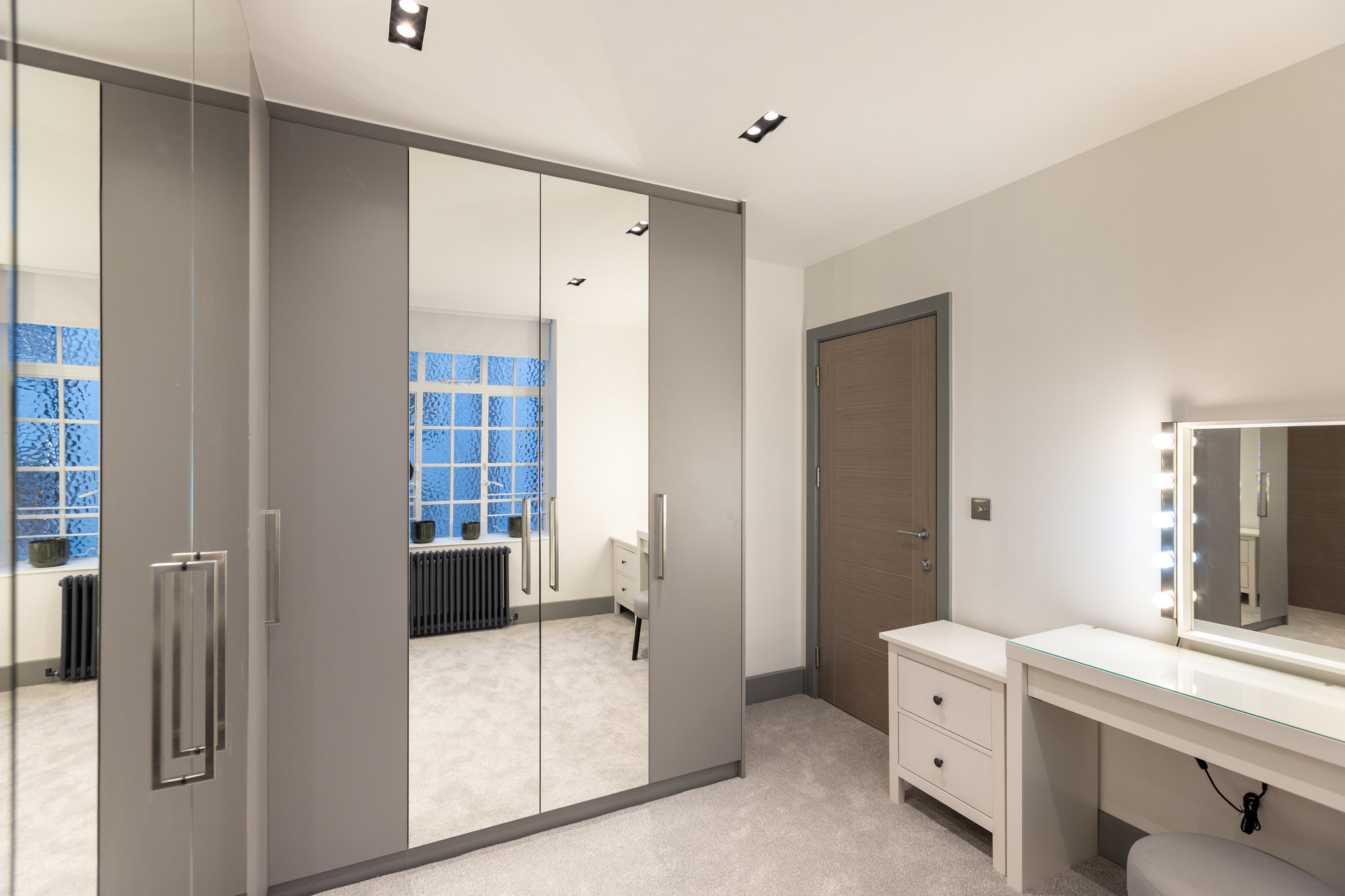 Bespoke walk-in wardrobe featuring grey panel and mirrored doors with vanity table and white dressing unit in Kensington