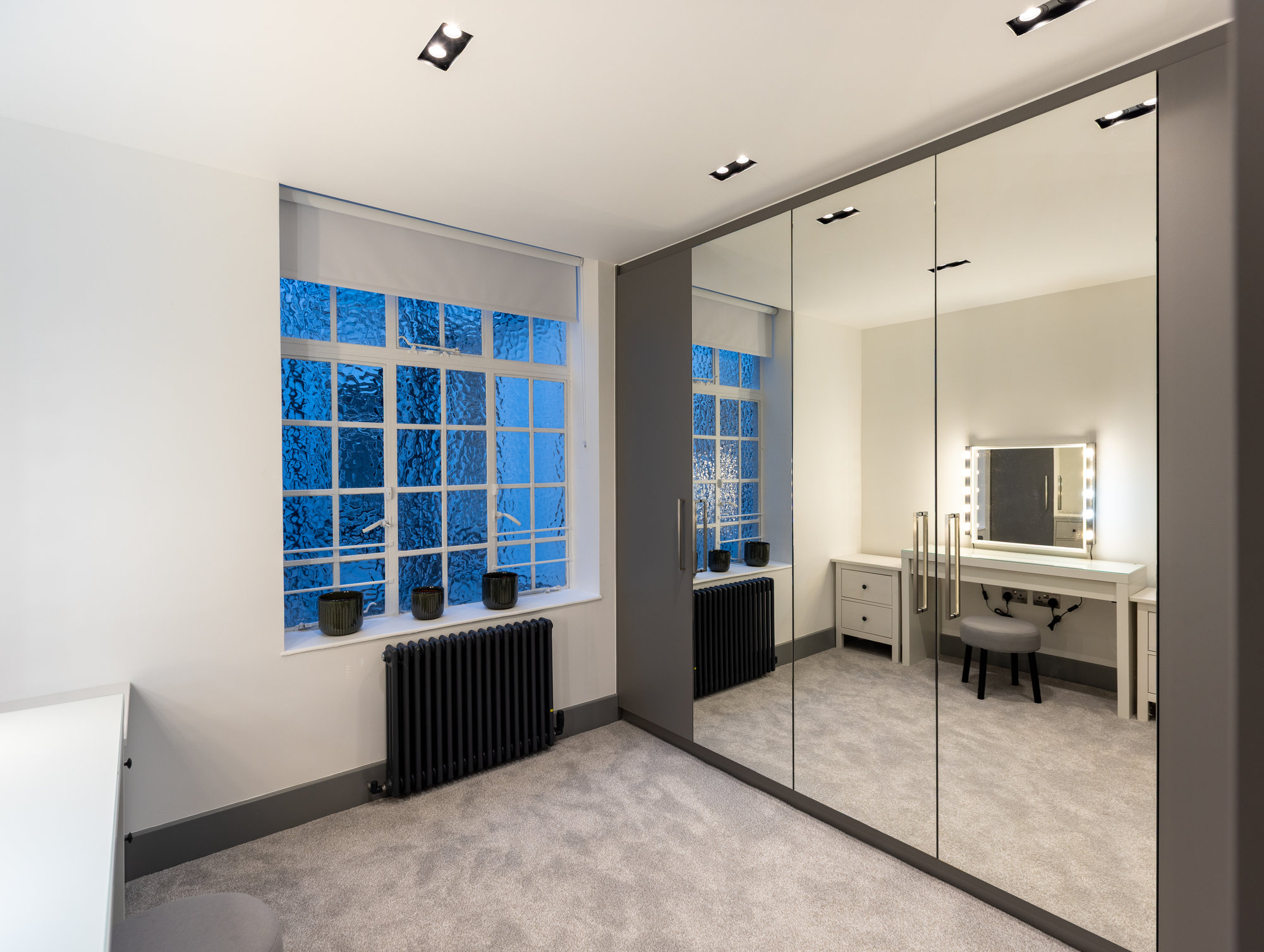 Bespoke walk-in wardrobe with mirrored and grey panel doors alongside a frosted window and vanity area in Kensington