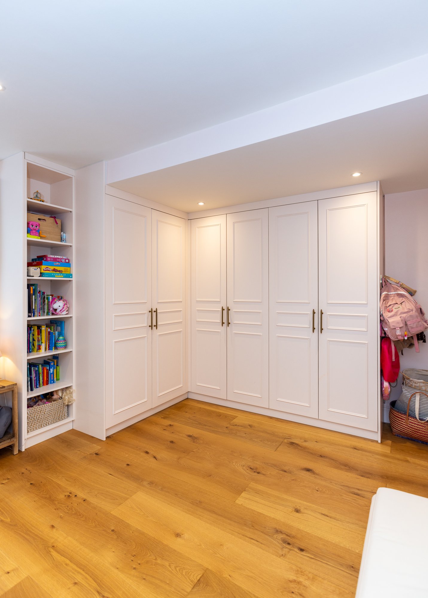 Wide-angle view of bespoke white corner fitted wardrobes with panel doors and integrated open shelving beside a children's bed in a Kensington home