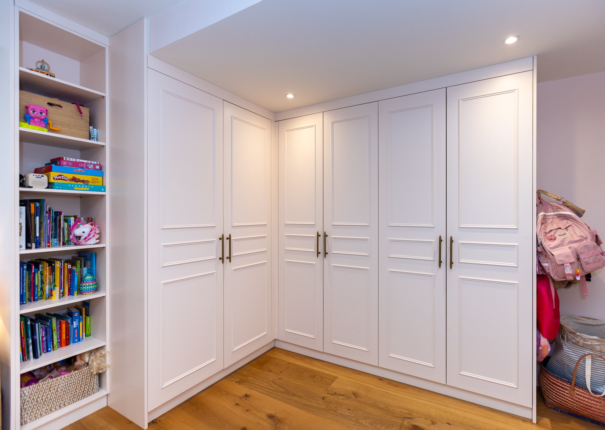 Bespoke white L-shaped corner fitted wardrobes with panel doors, antique brass handles, and integrated open bookshelf in a Kensington children's bedroom