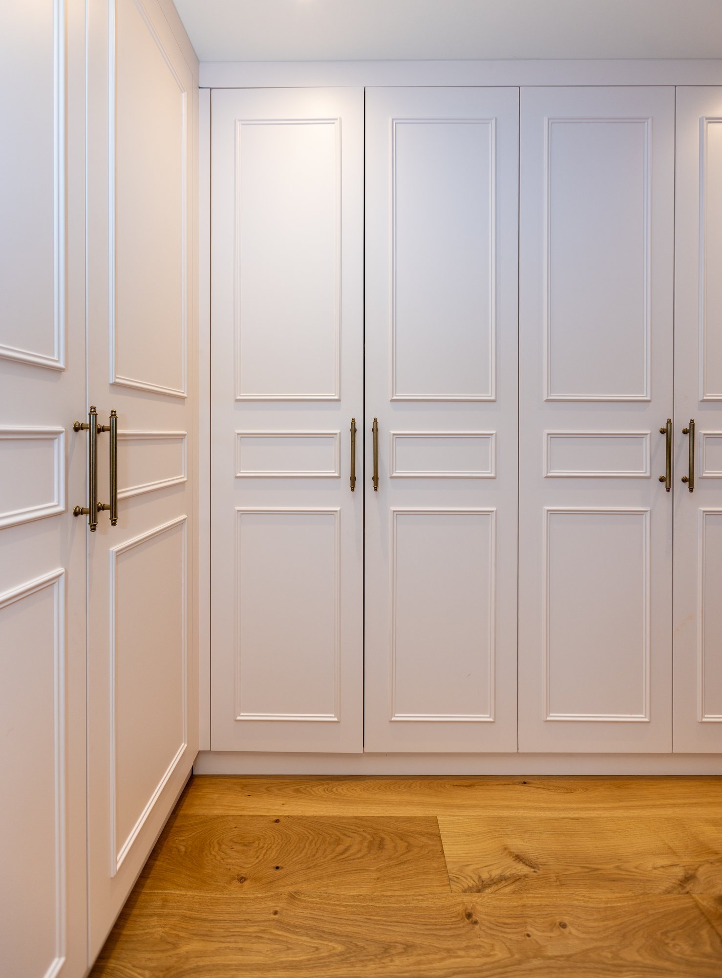Bespoke white L-shaped corner fitted wardrobes with panel doors, antique brass handles, and integrated open bookshelf in a Kensington children's bedroom