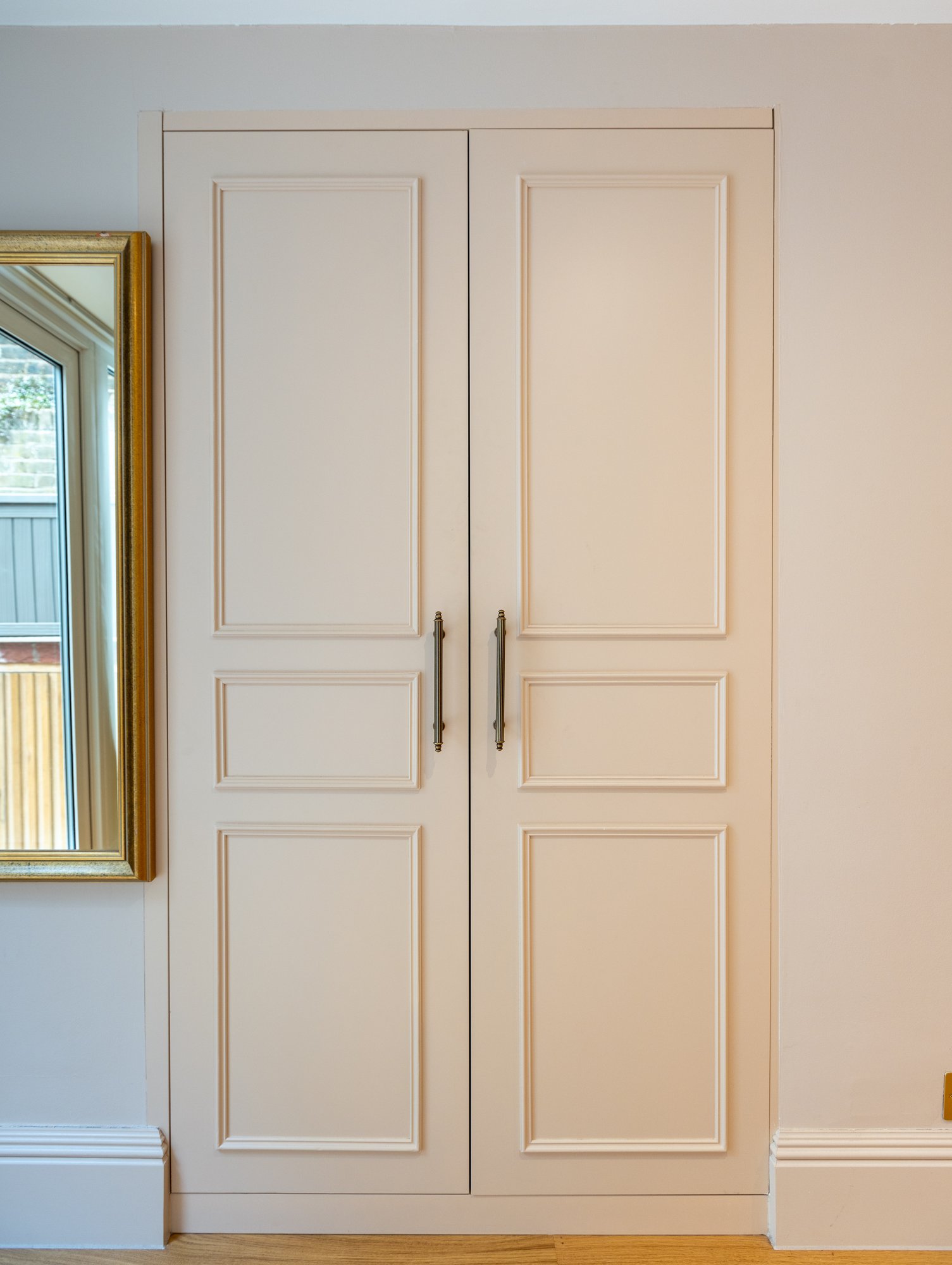 Bespoke white fitted wardrobe with three-panel shaker-style doors and antique brass bar handles in a Kensington bedroom