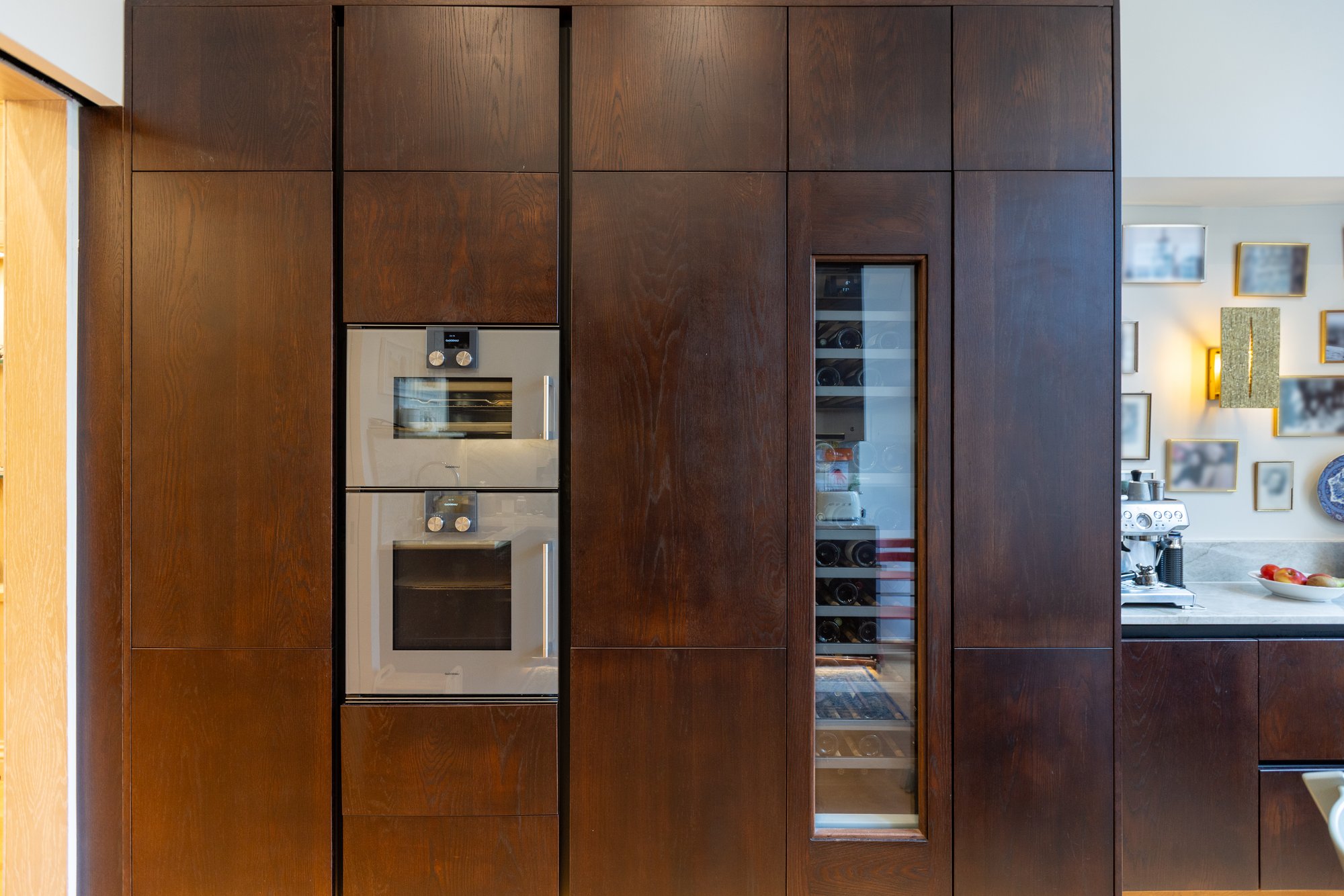 Bespoke dark oak kitchen appliance tower with integrated double ovens and glass-fronted wine cooler column in a Kensington home