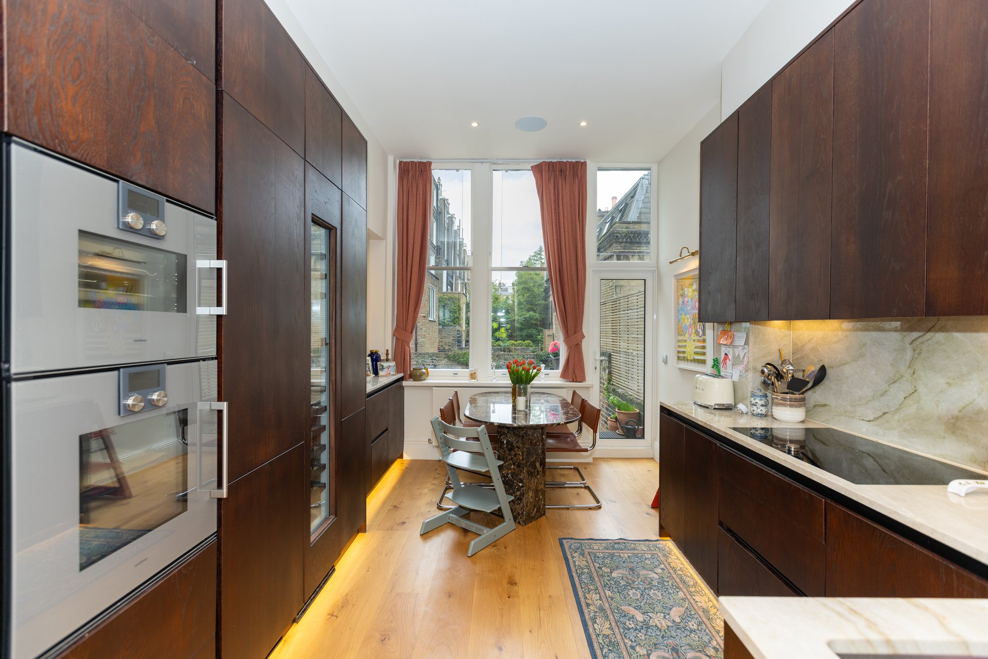 Full galley view of bespoke dark oak kitchen with marble worktop and splashback, integrated appliances, under-cabinet LED lighting, and dining area beyond in Kensington