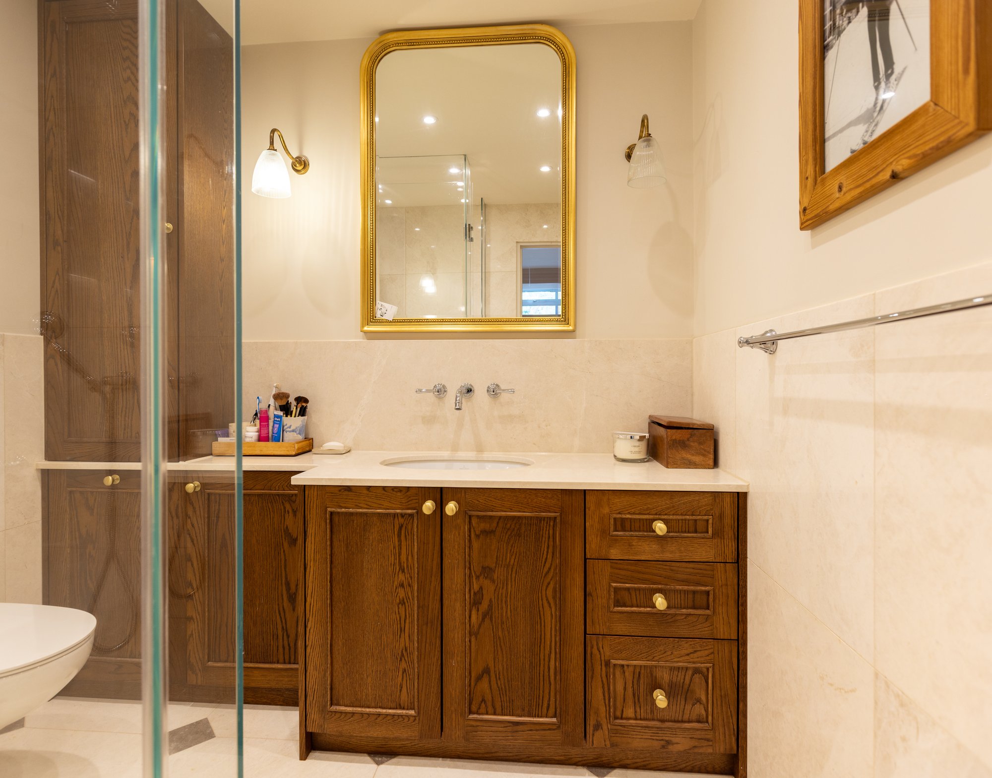 Bespoke dark oak bathroom vanity unit with marble worktop, wall-mounted chrome taps, and a gold-framed mirror with twin brass sconces in a Kensington home