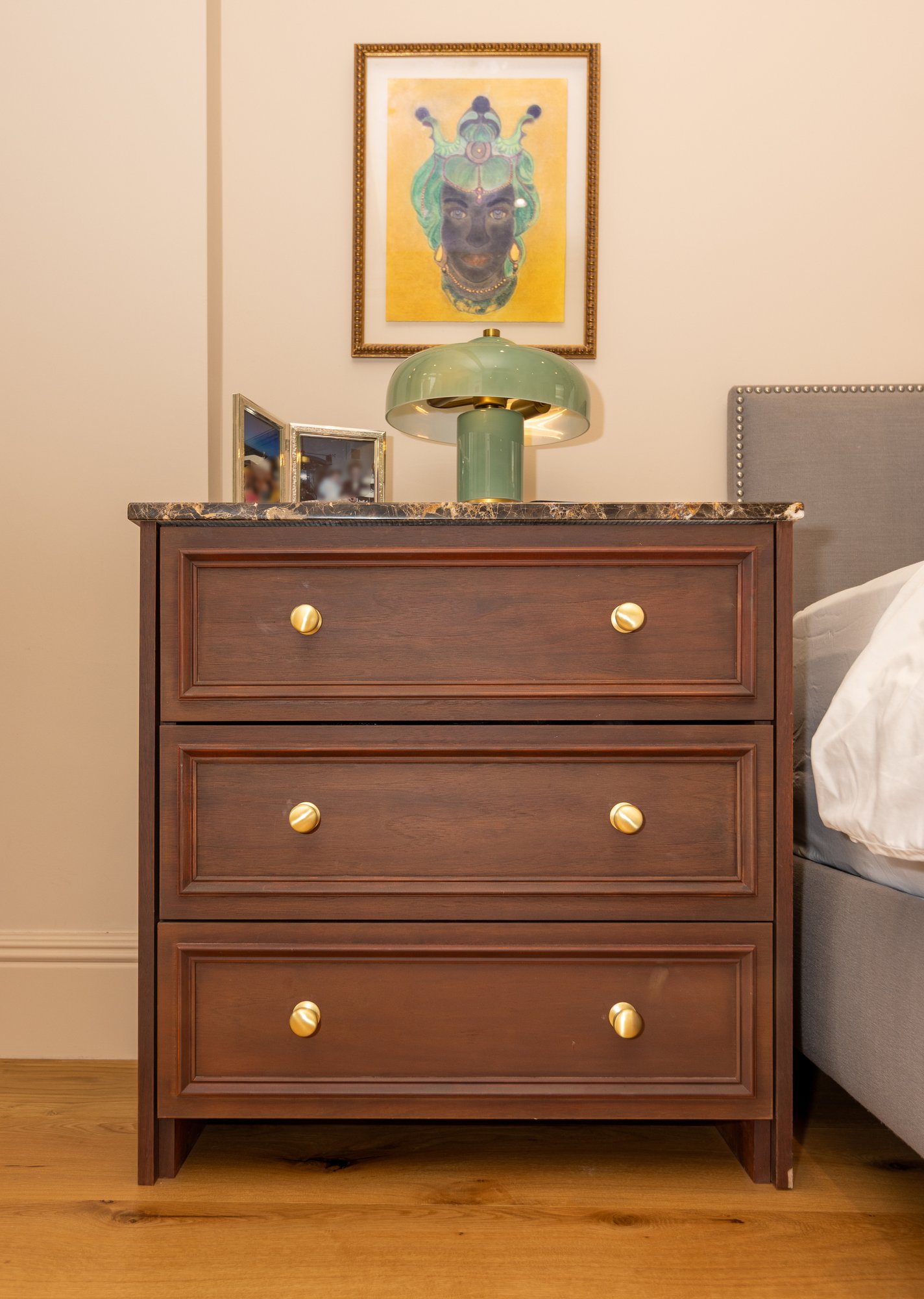 Bespoke dark oak bedside chest of drawers with marble top and brass knob handles beside an upholstered bed in a Kensington bedroom